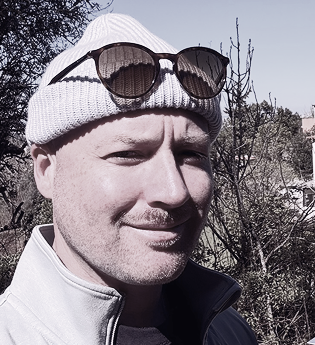 Portrait of Joseph Frederick Flynn, Founder and CEO of Ozeaon, wearing a white beanie with sunglasses pushed up, outdoors among bare trees.